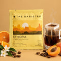 Lamora The Baristro Ethiopia Yirgacheffe drip bag coffee with fresh orange, apricot, and coffee beans on a tan table.